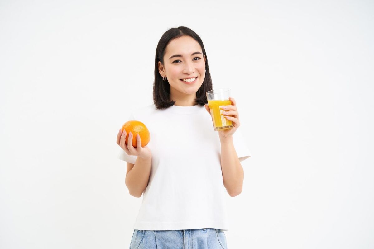 Perempuan memegang segelas jus jeruk dan sebuah jeruk di masing-masing tangannya