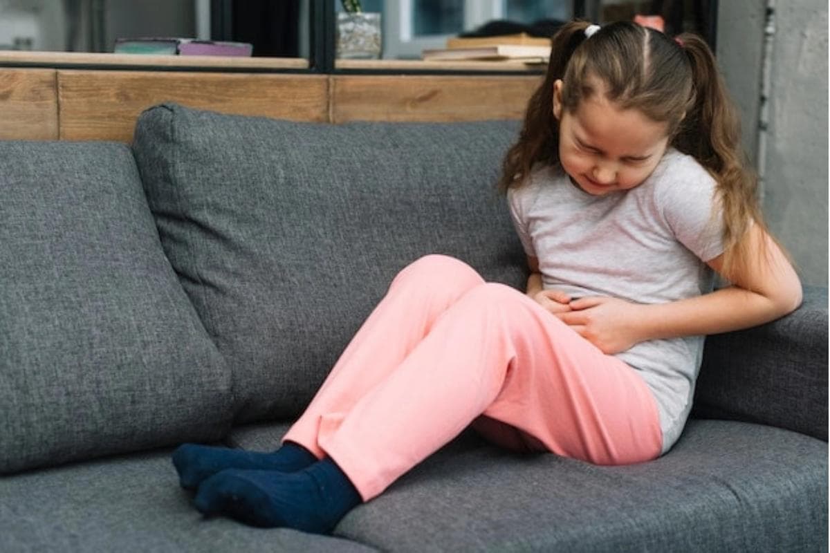 Anak perempuan duduk di sofa kesakitan memegangi perutnya
